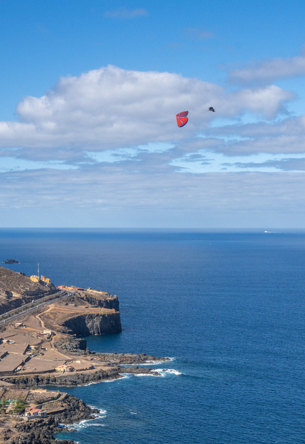 Christian Fernández del Valle realizando una maniobra acrobática en parapente biplaza sobre el mar, con el cielo despejado y las olas visibles en el fondo.