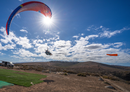 Vuelo en biplaza sobre el despegue de Los Giles en Gran Canaria, con un parapente rojo y la montaña de fondo bajo un cielo despejado.